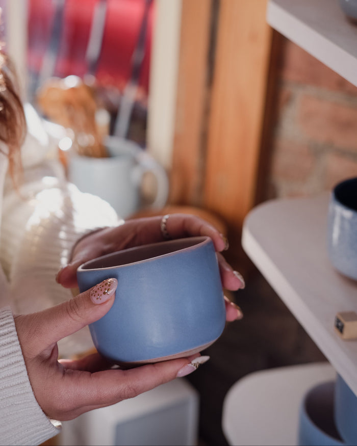 A woman holding a light baby blue ceramic mug with both hands, set against a warm, softly blurred background of shelves and ceramic home decor.