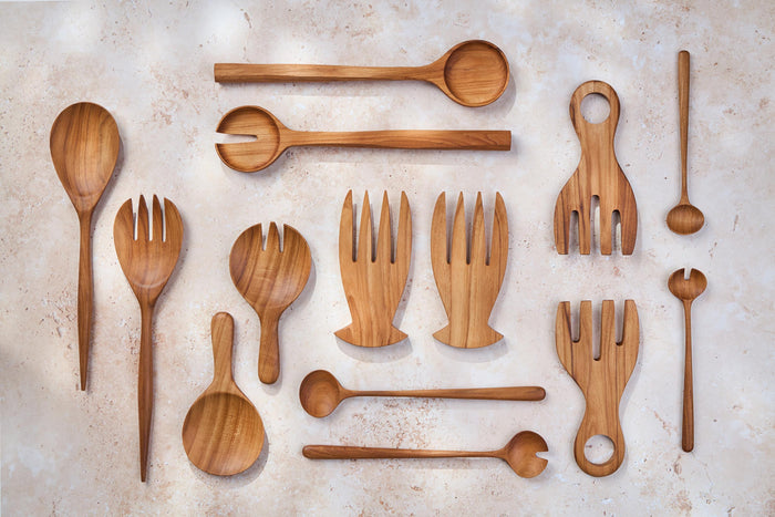 A variety of Palinopsia wooden teak spoons, forks, and salad serving sets in many shapes and sizes on a light stone background.