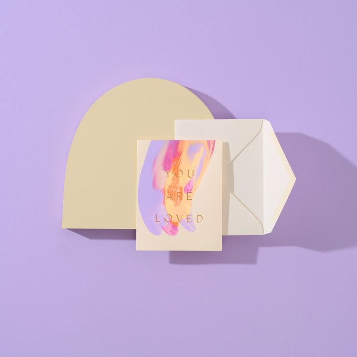 You Are Loved handmade purple artisanal greeting card with purple, yellow and pink paint swirls and an envelope on a purple geometric background.