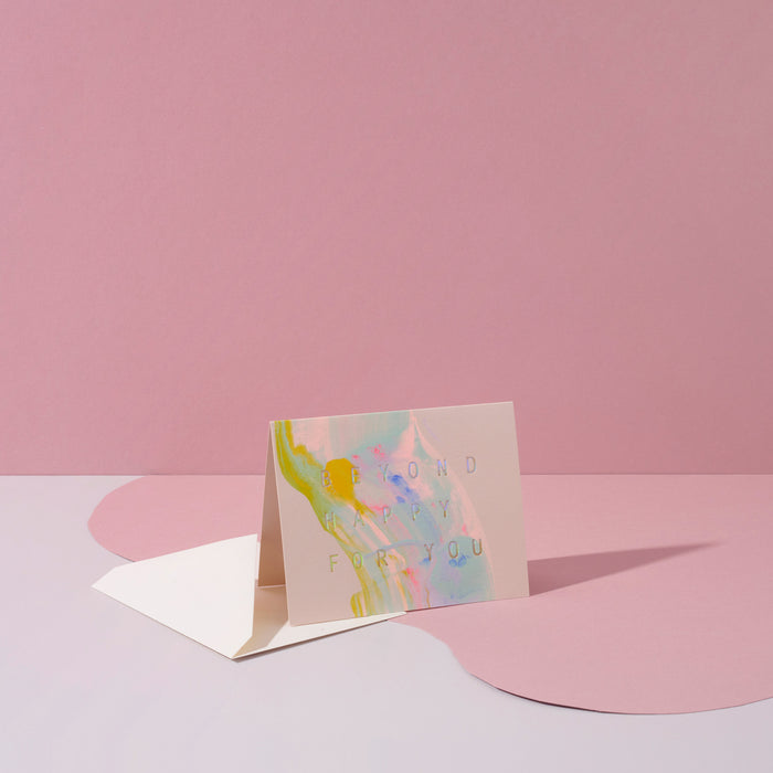 Beyond Happy For You handmade artisanal greeting card with blue, pink and yellow paint swirls and an envelope on a pink geometric background.