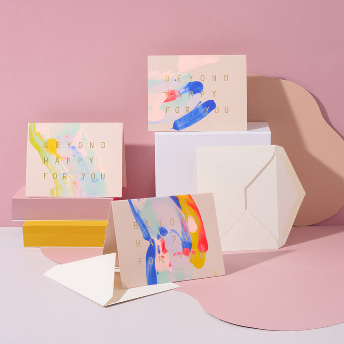 Three Beyond Happy For You handmade artisanal greeting cards with blue, pink and yellow paint swirls with envelopes on a pink geometric background.