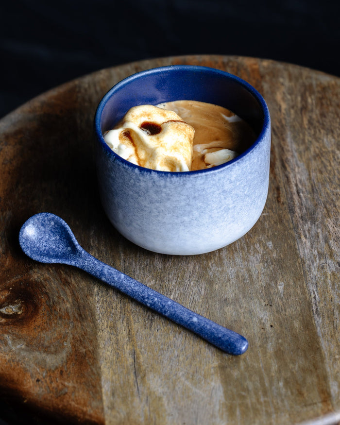 Dark and moody photo of a handmade ceramics cup and teaspoon in dark blue and white resembling a snow blizzard with affogatto on a bar stool.