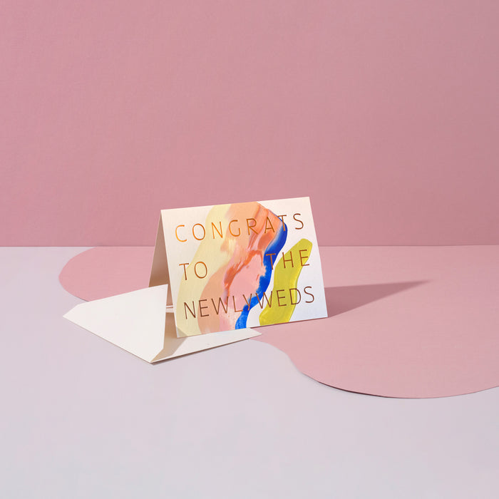 Congrats To The Newlyweds handmade purple artisanal greeting card with, pink and yellow paint swirls and an envelope on a pink geometric background.