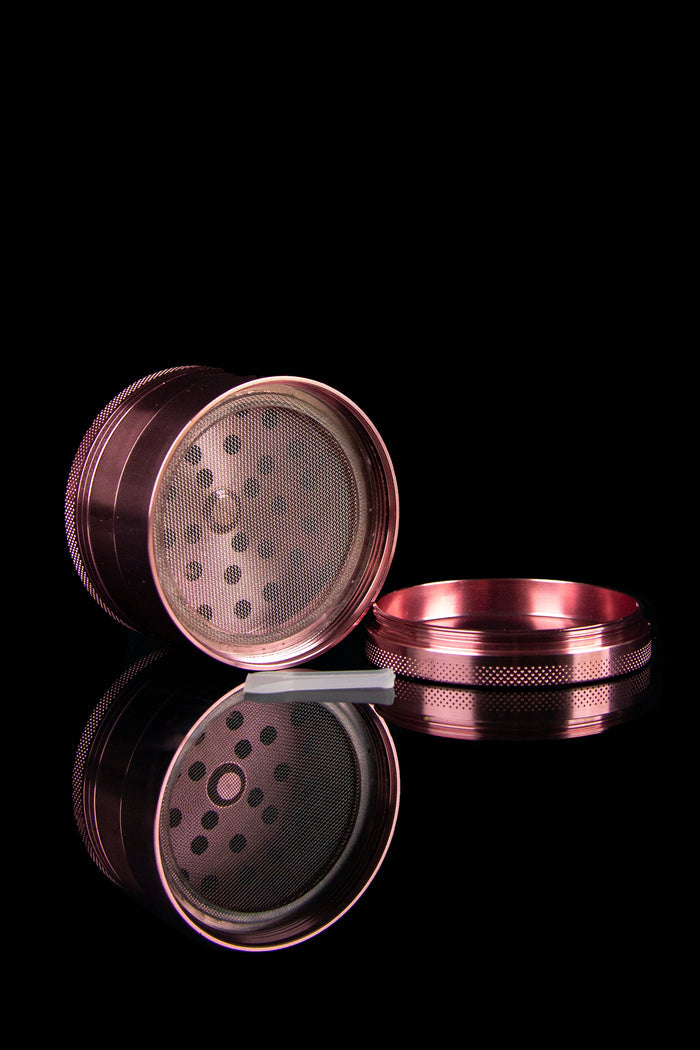 Disassembled pink metal cannabis grinder with mesh screens and grinding teeth visible against a black background.