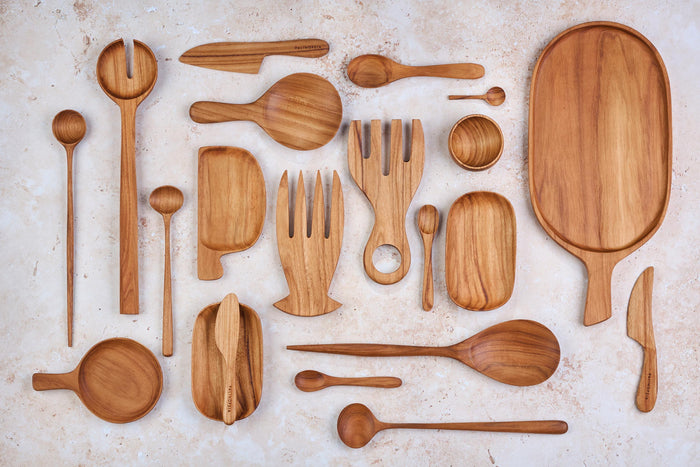 A variety of Palinopsia wooden teak serving dishes, salad servers, bowls, trays, knives, cheese boards in many shapes and sizes on a light stone background.