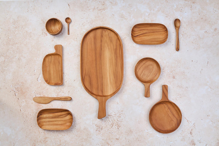 A mix of Palinopsia wooden teak serving trays, knives, and cheese boards on a light stone background.