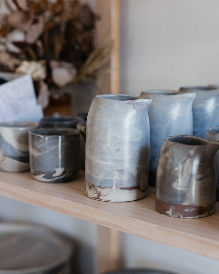 Wooden shelf in a shop with ceramic handmade pitchers and coffee cups, out of focus dried flowers decorate the background