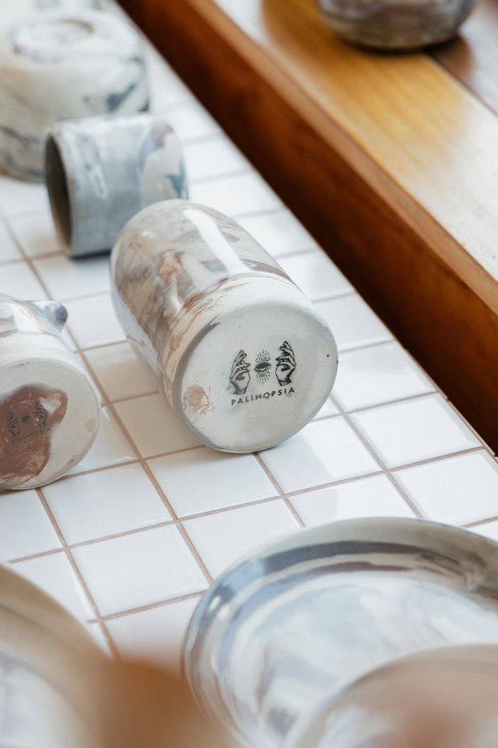 Tiled shelf in a shop with a ceramic handmade pitcher on its side displaying the Palinopsia logo, marbled neutral clay bowls, plates and mugs are placed around