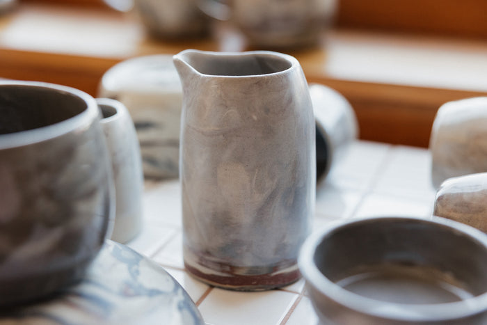 Tiled shelf in a shop with a ceramic handmade pitcher in focus, marbled neutral clay bowls decorate the foreground
