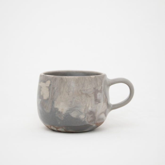 A handcrafted ceramic mug with a marbled pattern in shades of gray, with a handle on the right side, on a white background.