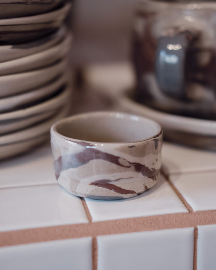 A handmade ceramic small bowl with a marbled pattern of brown, beige, and white, displayed on a white tiled surface with other ceramic dishes blurred in the background.