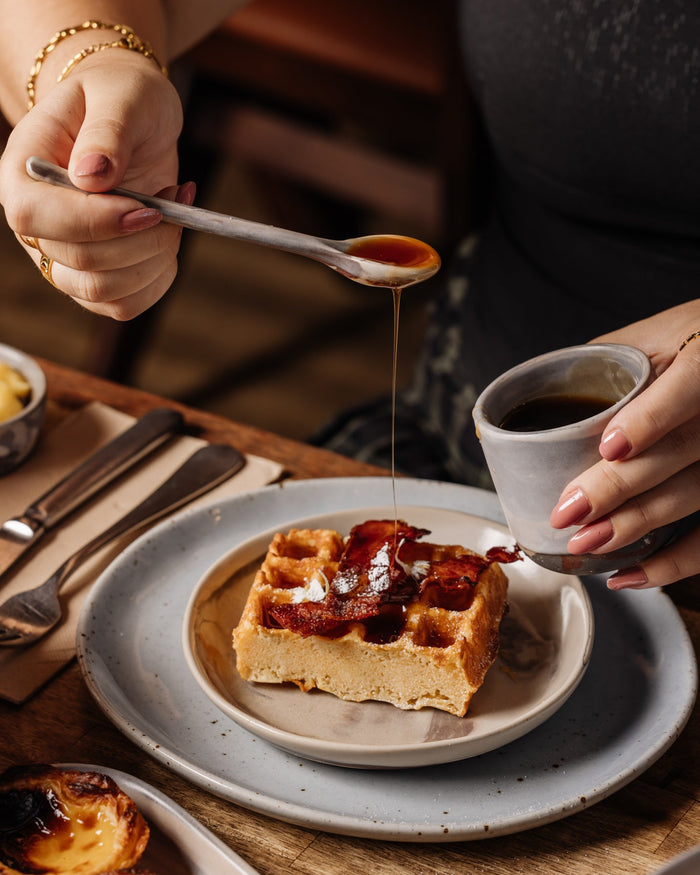 Person pouring syrup onto a waffle with bacon and with a ceramic holding a handmade espresso cup