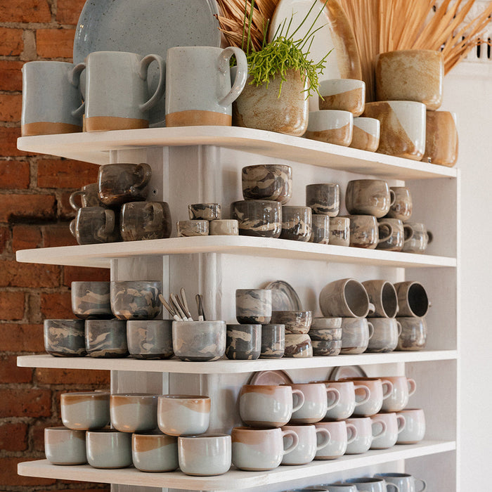 A variety of handmade ceramic pitchers, plates, salt pinches, mugs, espresso cups, tumbler cups and teaspoons on display in blue, marbled clays, pink, white, and orange.