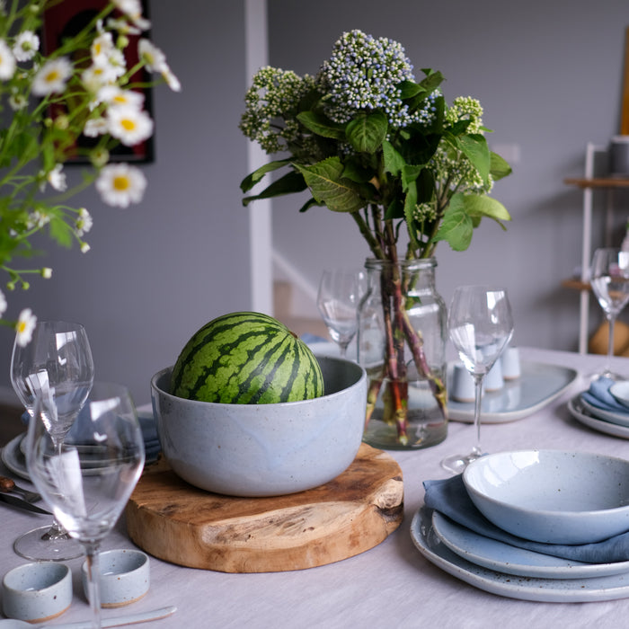 Blue grey dinnerware setting by Palinopsia Ceramics with fresh flowers, watermelon and a grey tablecloth