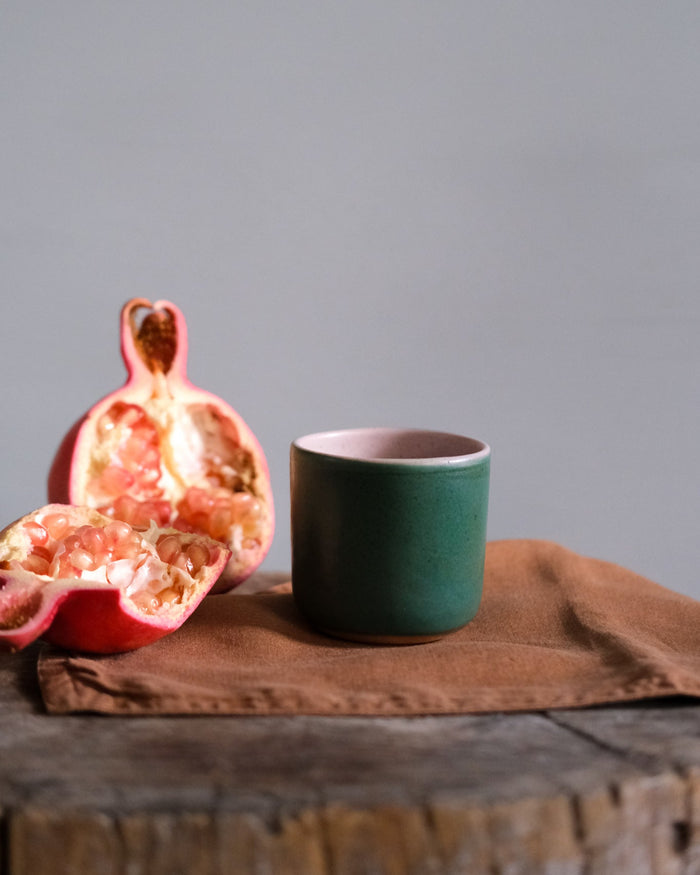 Green stoneware cup on a brown napkin with a halved pomegranate on a wooden surface.