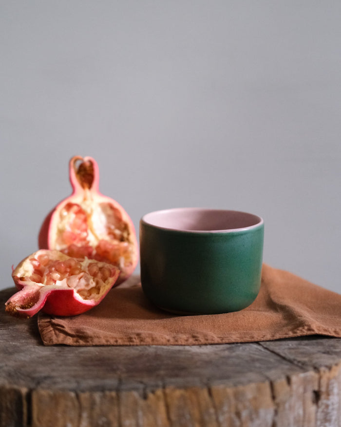 Green ceramic mug with pink interior on a brown napkin with a pomegranate on a wooden surface