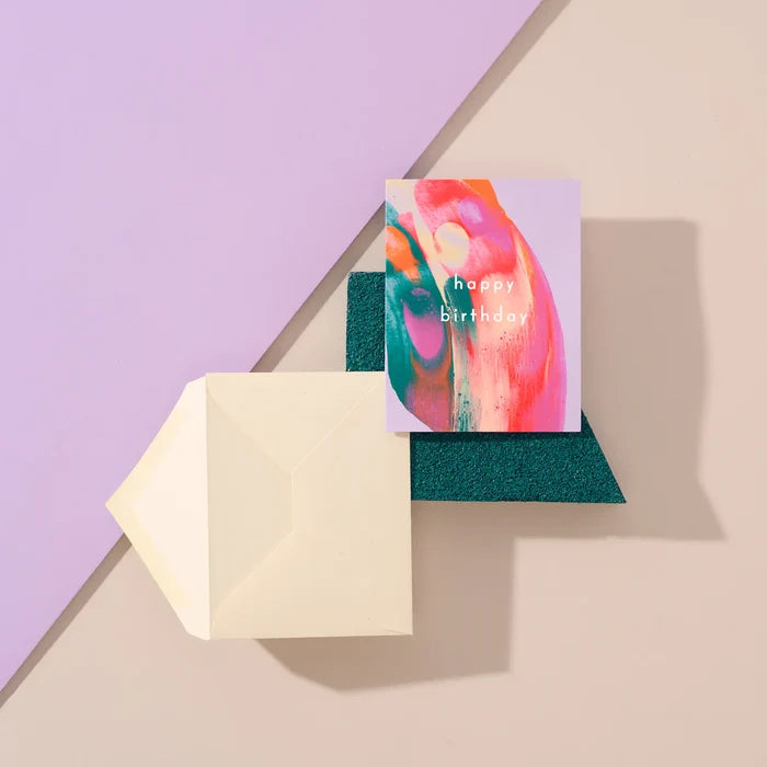 Happy Birthday handmade purple artisanal greeting card with green and pink paint swirls and an envelope on a purple geometric background.