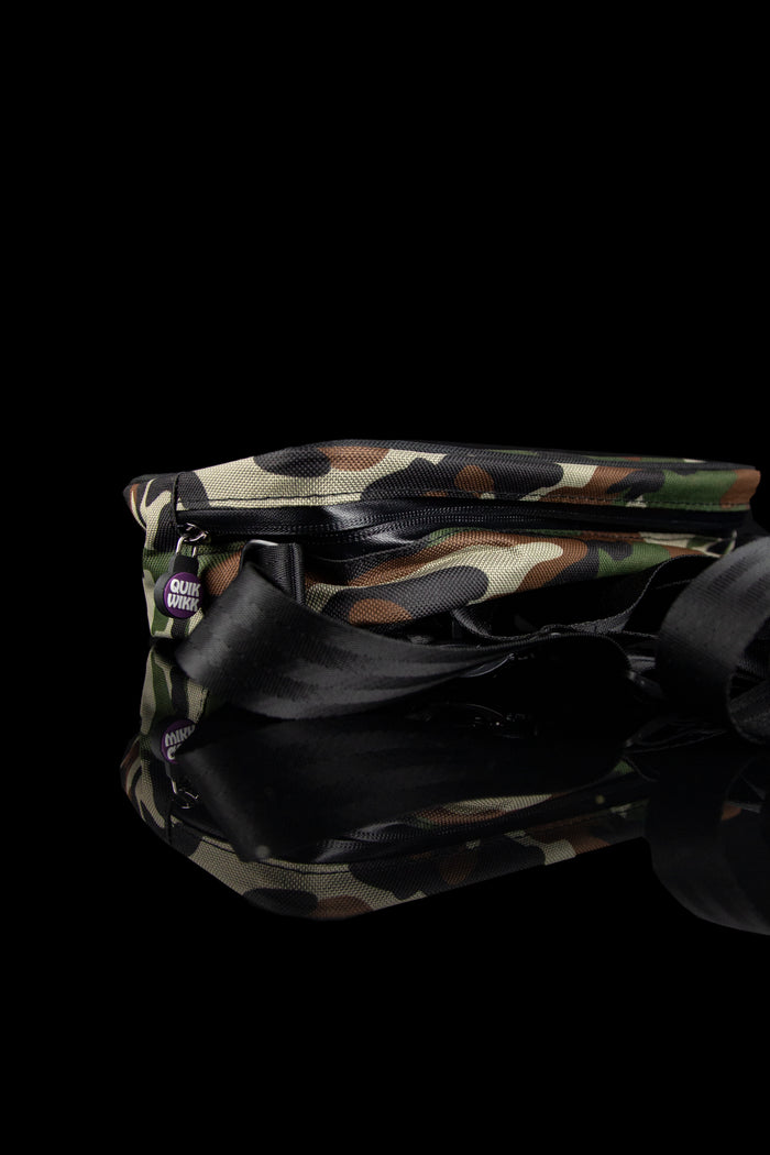Low-angle close-up of a camo Quik Wikk Chest Pack showing the textured top, zipper and reflective black strap against a black background
