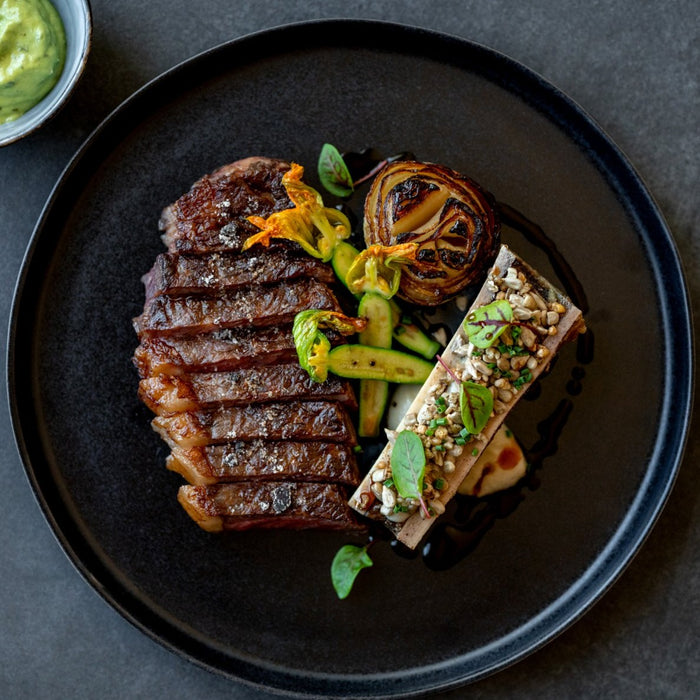 Birds-eye view of a handmade black ceramic dinner plate by Palinopsia Ceramics served with perfectly grilled steak, bone marrow and grilled vegetables by Sydney chef Gerald Touchard from the Northern Beaches