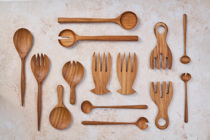 A variety of Palinopsia wooden teak spoons, forks, and salad serving sets in many shapes and sizes on a light stone background.